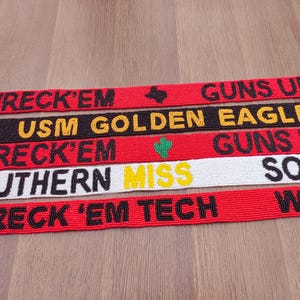 May include: Several beaded belts in red, black, and white are displayed. The belts have text in black and yellow, with phrases such as "WRECK 'EM", "GUNS UP", and "SOUTHERN MISS". Small graphic details are also present.