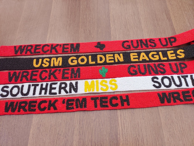 May include: Several beaded belts in red, black, and white are displayed. The belts have text in black and yellow, with phrases such as "WRECK 'EM", "GUNS UP", and "SOUTHERN MISS". Small graphic details are also present.