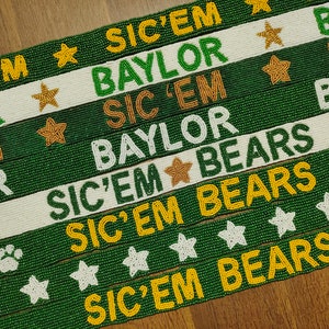 May include: Green beaded belts with the words "SIC'EM", "BAYLOR", and "BEARS" in gold and white. Some belts feature star and paw print designs. The belts are displayed on a wooden surface.