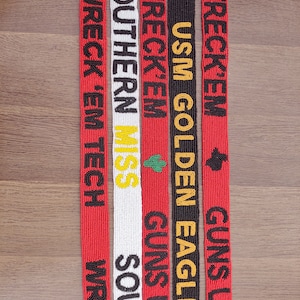 May include: A selection of beaded belts with text in various colours. The belts are red, white, and black. Text includes "WRECK 'EM TECH", "SOUTHERN MISS", and "USM GOLDEN EAGLES". Each belt has a unique design and text arrangement.