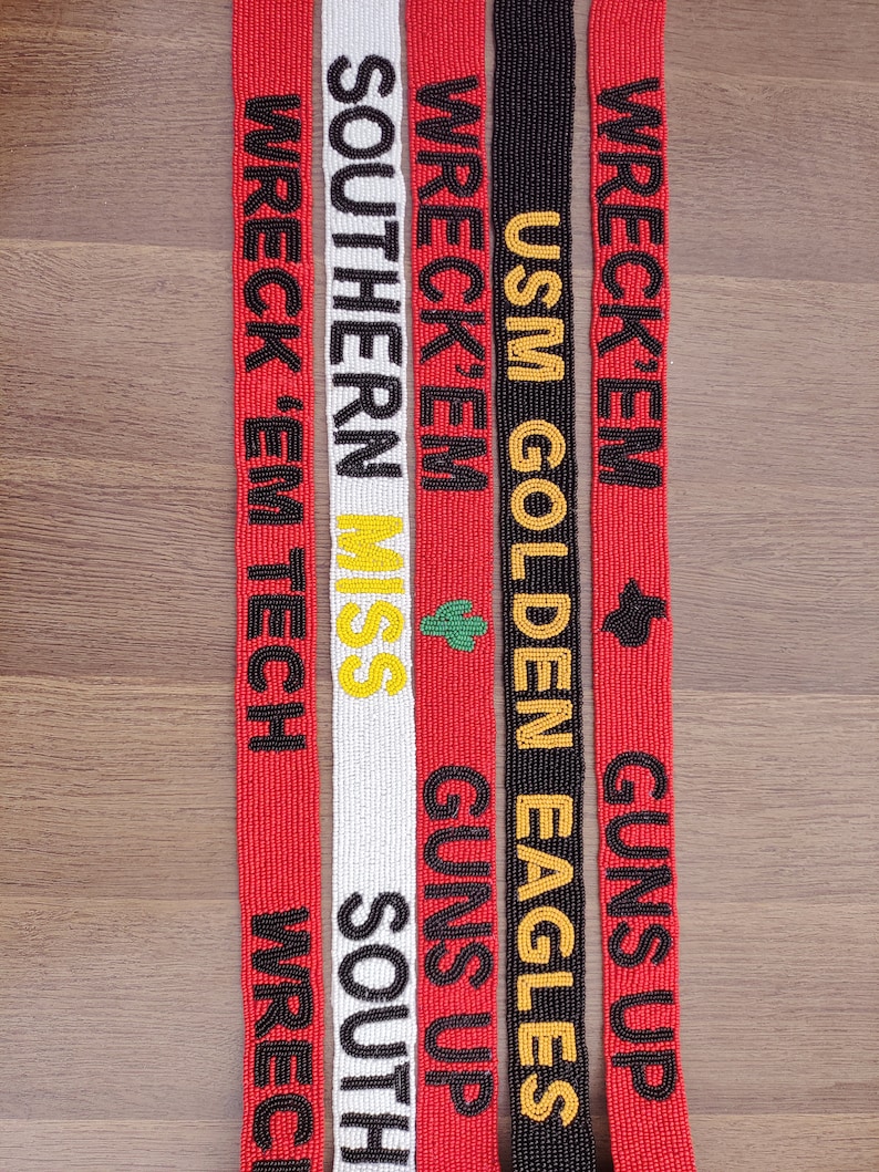 May include: A selection of beaded belts with text in various colours. The belts are red, white, and black. Text includes "WRECK 'EM TECH", "SOUTHERN MISS", and "USM GOLDEN EAGLES". Each belt has a unique design and text arrangement.