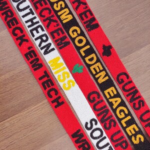 May include: Close-up of several beaded lanyards with text. The lanyards are red, black, and white, with phrases like "Wreck 'Em," "Guns Up," and "USM Golden Eagles" in various colours. The lanyards are on a wooden surface.