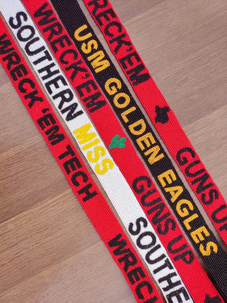 May include: Close-up of several beaded lanyards with text. The lanyards are red, black, and white, with phrases like "Wreck 'Em," "Guns Up," and "USM Golden Eagles" in various colours. The lanyards are on a wooden surface.