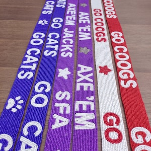 May include: A collection of beaded lanyards in various colors, including purple, blue, white, and red. Each lanyard features white lettering and decorative elements like paw prints and stars, with phrases like "GO CATS" and "GO COOGS".