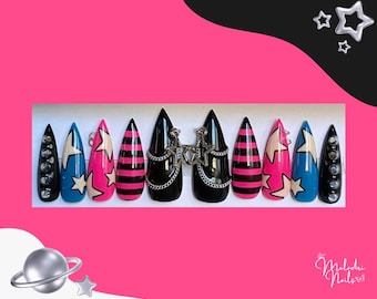 Puede incluir: Un conjunto de uñas postizas con forma de estilete en varios diseños. Las uñas presentan bases negras, rosas y azules con patrones de estrellas y rayas, detalles de cadena plateada y detalles con tachuelas. El conjunto incluye una variedad de colores y adornos.