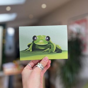 Peut inclure: Une carte peinte à la main représentant une grenouille verte aux grands yeux noirs. La grenouille est assise sur une feuille verte.