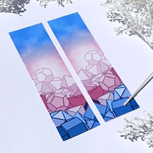 Può includere: Due segnalibri con un design geometrico di cristalli rosa, blu e bianchi su uno sfondo sfumato blu e rosa.