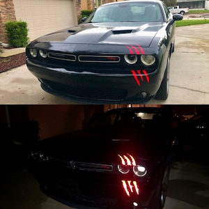 May include: Black Dodge Challenger with red claw mark decals on the front bumper and hood. The car has a black grille and the word "Challenger" is visible on the front. The car is parked on a driveway.