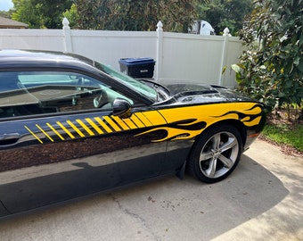 May include: A black car with yellow and black racing stripes and flames on the side. The car is parked on a driveway.