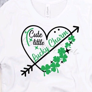 Puede incluir: Camiseta blanca con un diseño de corazón negro que dice "Cute little lucky charm" con una flecha negra y tréboles verdes.