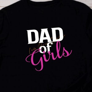 Może przedstawiać: Czarny T-shirt z napisem "DAD of GIRLS" w białym i różowym kolorze.