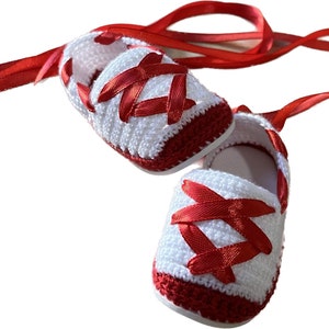 May include: A pair of white crocheted baby shoes with red satin ribbon laces. The shoes have a red trim around the edges.