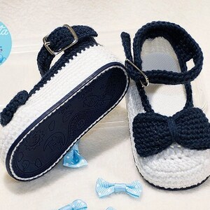May include: White and navy blue crochet baby shoes with a strap and bow detail. The soles are navy blue with a pattern. Includes three small light blue bows.