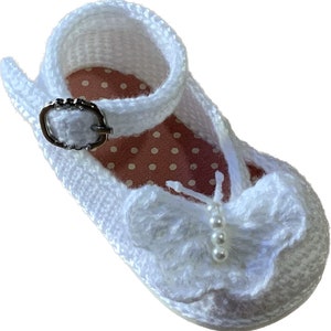 May include: White crocheted baby shoes with a white butterfly bow and pearl accents. The shoes have a white strap with a silver buckle.