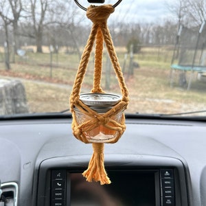 May include: A yellow rope car air freshener with a glass jar filled with a light orange substance. The air freshener is hanging from the rearview mirror of a car.