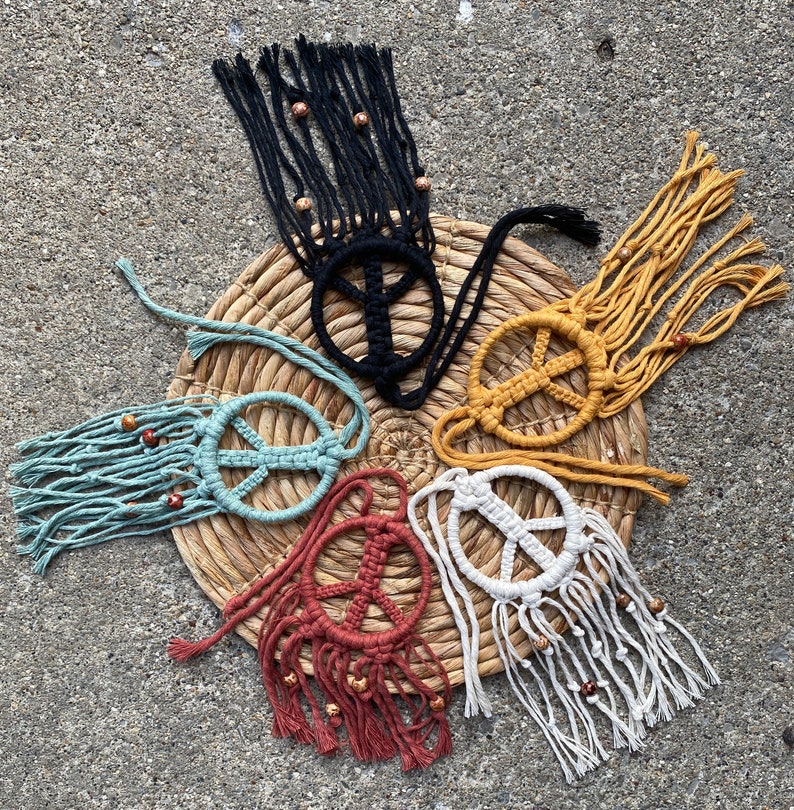 Peut inclure: Six suspensions murales en macram&eacute; en forme de signe de paix avec des franges et des perles. Les signes de paix sont de diff&eacute;rentes couleurs, notamment le noir, le blanc, le jaune, le bleu et le rouge.