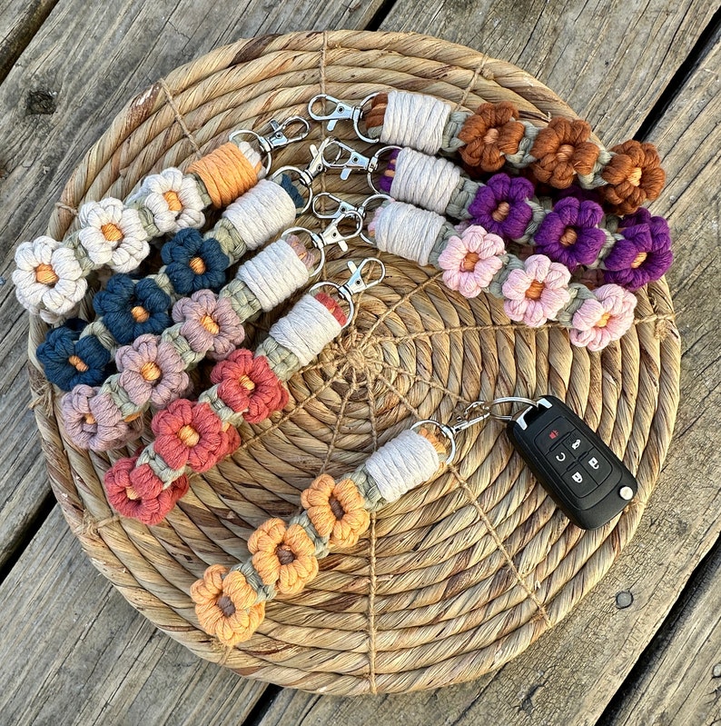 May include: A set of six colorful crocheted keychains with flower designs. Each keychain features a different color combination and a silver key ring. The keychains are displayed on a woven straw mat.