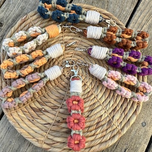 May include: A set of seven colorful macrame keychains with different patterns and colors, including orange, purple, brown, and white. The keychains are made with a thick, knotted cord and feature a silver key ring.