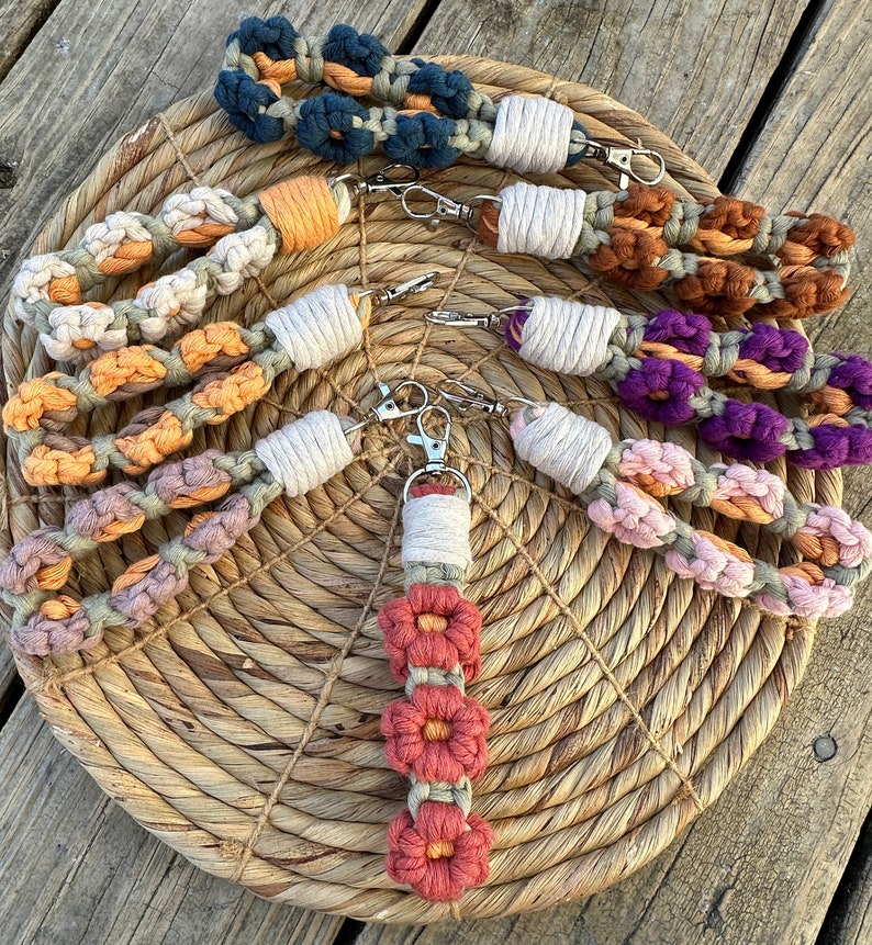 May include: A set of seven colorful macrame keychains with different patterns and colors, including orange, purple, brown, and white. The keychains are made with a thick, knotted cord and feature a silver key ring.