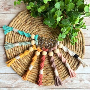 May include: A collection of handmade macrame keychains in various colors, including teal, yellow, and pink. Each keychain features a wooden bead and a metal clasp. The keychains are arranged on a woven mat with greenery in the background.