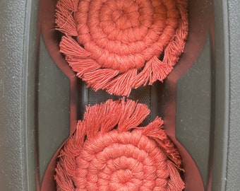 May include: Two red crocheted coasters with fringe, sitting in a black cup holder.