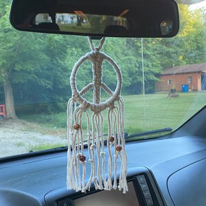 Peut inclure: Un symbole de paix en macram&eacute; blanc suspendu au r&eacute;troviseur d'une voiture. Le symbole de paix a des perles en bois qui pendent du bas.