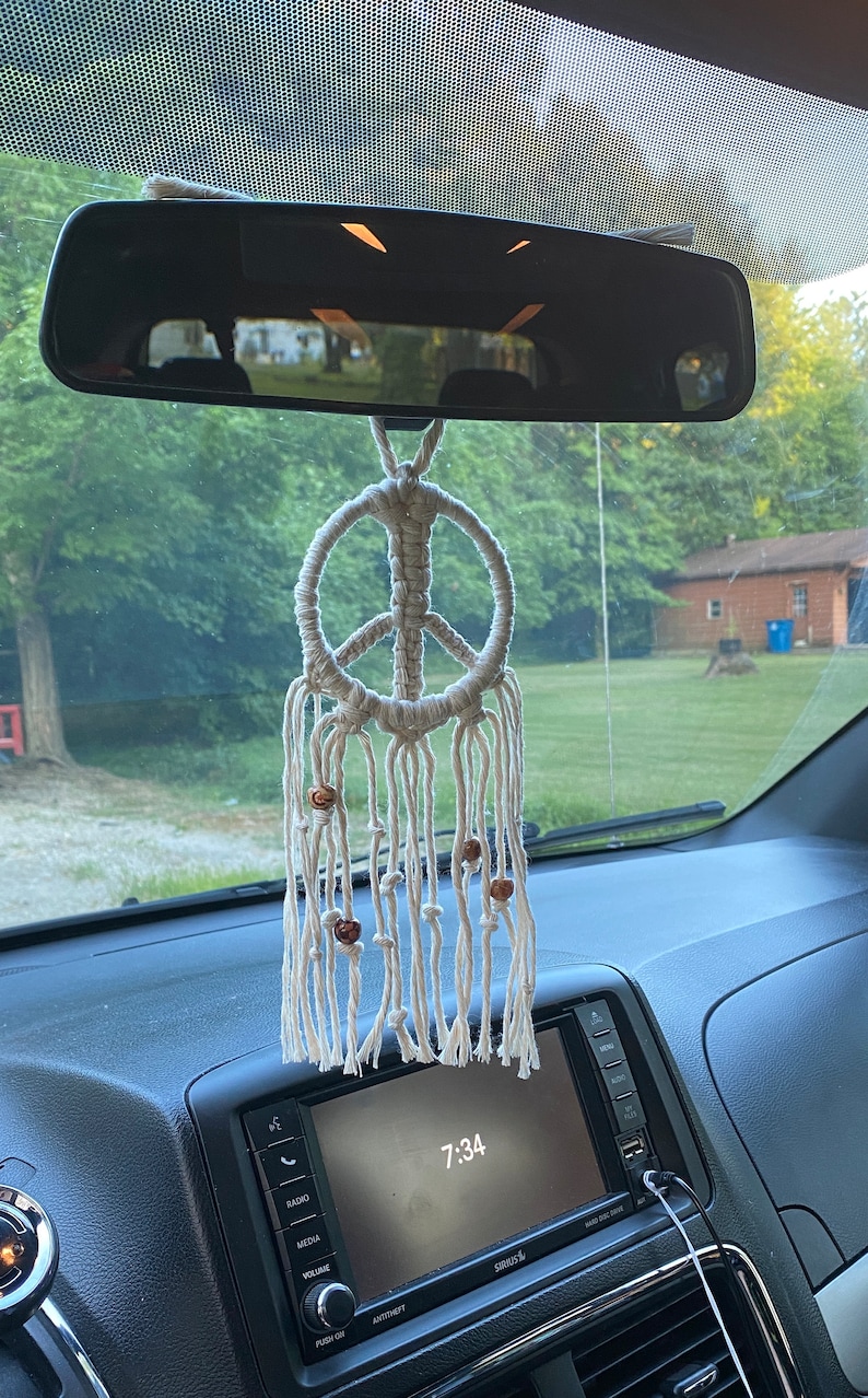 Peut inclure: Un symbole de paix en macram&eacute; blanc suspendu au r&eacute;troviseur d'une voiture. Le symbole de paix a des perles en bois qui pendent du bas.