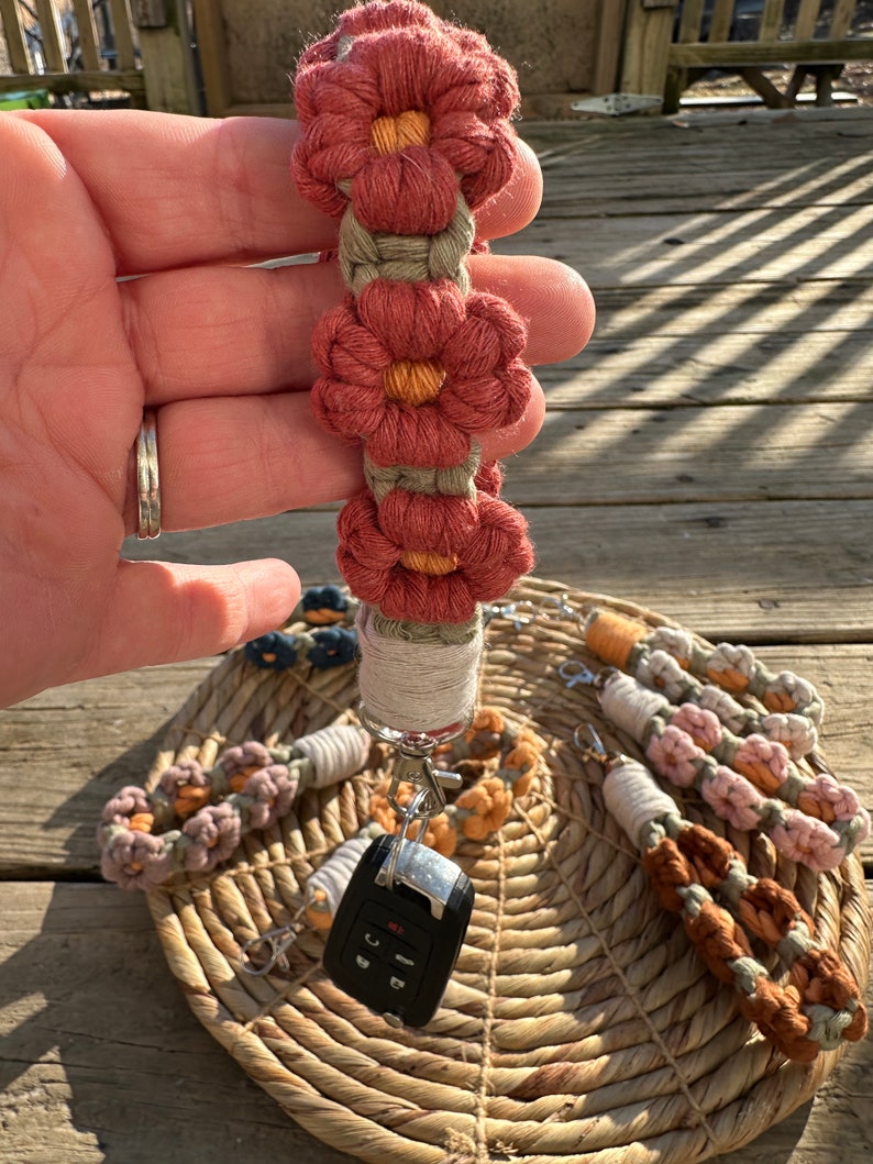 May include: A set of six colorful crocheted keychains with flower designs. The keychains are attached to a silver key ring and a black car key. The keychains are displayed on a woven basket.