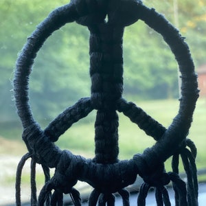 Peut inclure: Un symbole de paix en macram&eacute; noir avec de longues franges qui pendent du bas.