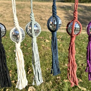 May include: Six macrame disco ball ornaments in black, cream, light green, navy, rust, and purple. Each ornament features a mirrored disco ball encased in a macrame net, with a matching tassel. The ornaments are hanging outdoors.