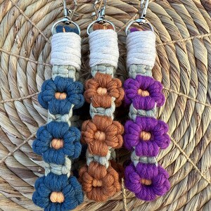 May include: Three crocheted keychains with different colored flowers. The keychain on the left has blue flowers, the middle keychain has brown flowers, and the keychain on the right has purple flowers.