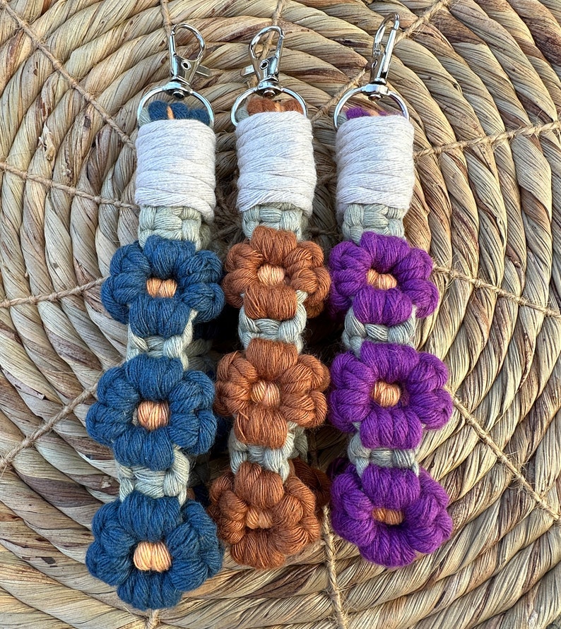 May include: Three crocheted keychains with different colored flowers. The keychain on the left has blue flowers, the middle keychain has brown flowers, and the keychain on the right has purple flowers.