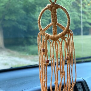 Peut inclure: Un porte-bonheur de voiture en macram&eacute; en forme de signe de paix avec des perles brunes qui pendent du bas. Le porte-bonheur est suspendu au r&eacute;troviseur d'une voiture.