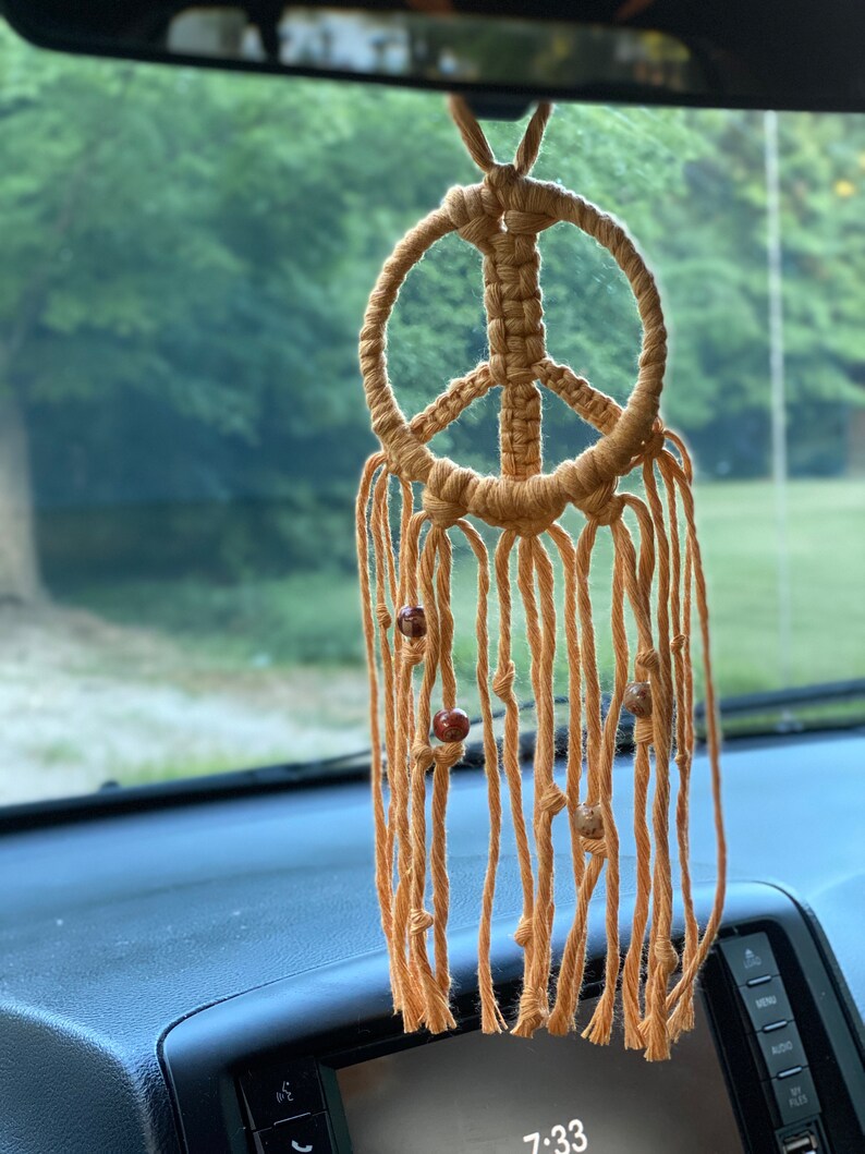Peut inclure: Un porte-bonheur de voiture en macram&eacute; en forme de signe de paix avec des perles brunes qui pendent du bas. Le porte-bonheur est suspendu au r&eacute;troviseur d'une voiture.
