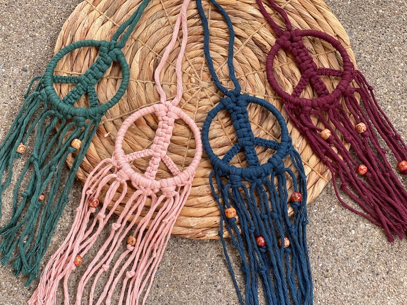 Peut inclure: Quatre tentures murales en macram&eacute; en forme de signe de paix avec des perles en bois. Les tentures sont en vert, rose, bleu et bordeaux.