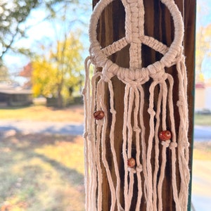 Peut inclure: Une suspension murale en macram&eacute; en forme de signe de paix blanc avec des perles en bois brun.
