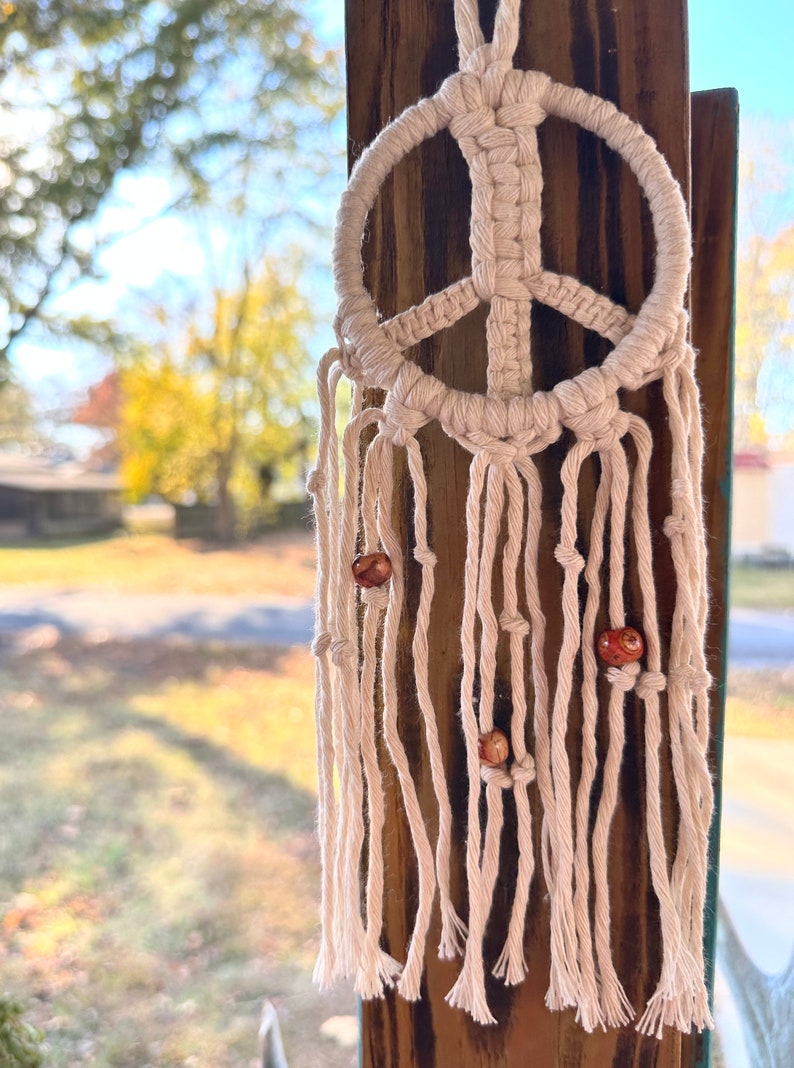 Peut inclure: Une suspension murale en macram&eacute; en forme de signe de paix blanc avec des perles en bois brun.