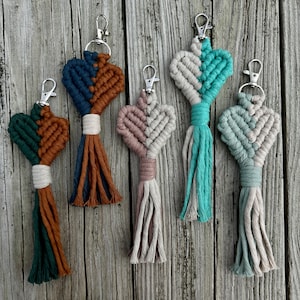 May include: Macrame heart-shaped keychains in various colors, including teal, green, and pink, with silver-tone clasps and tassel details. The keychains are arranged on a wooden surface.
