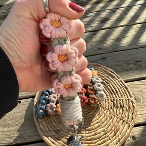 May include: A camera strap with a crocheted flower design in pink, orange, and green. The strap has a metal clip and is attached to a woven basket.