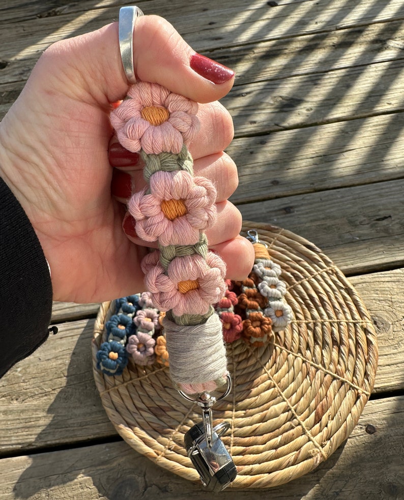 May include: A camera strap with a crocheted flower design in pink, orange, and green. The strap has a metal clip and is attached to a woven basket.