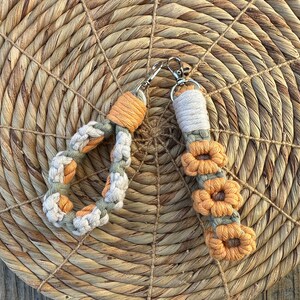 May include: Two macrame keychains with a flower design. One keychain is a circle with a light brown and green color scheme. The other keychain is a long strand with a light brown and orange color scheme.