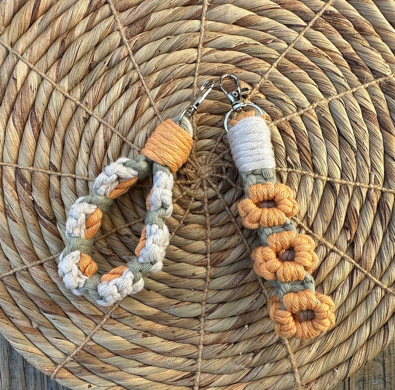 May include: Two macrame keychains with a flower design. One keychain is a circle with a light brown and green color scheme. The other keychain is a long strand with a light brown and orange color scheme.