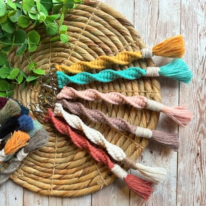 May include: A collection of macrame keychains in various colors, including mustard yellow, teal, and dusty rose. Each keychain features a twisted rope design and a tassel at the end. The keychains are displayed on a woven mat.