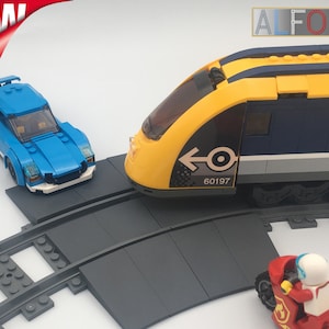 Può includere: Un'auto giocattolo blu e un treno giocattolo giallo e blu su un binario grigio. Il treno ha una freccia nera e il numero 60197 sul lato. Il treno è su un binario curvo.