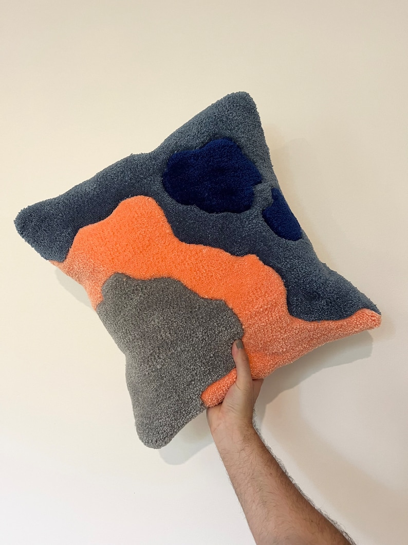 Op de afbeelding: Een vierkant kussen met een zachte, pluche textuur. Het kussen is gemaakt van drie kleuren: grijs, oranje en blauw. De kleuren zijn gerangschikt in een abstract patroon.