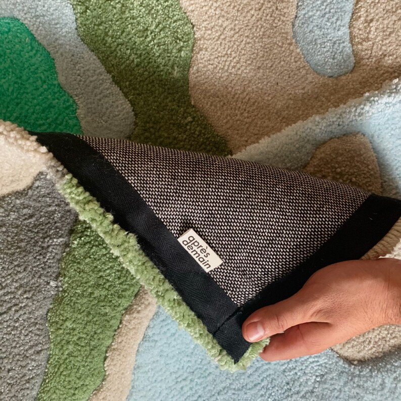 K&ouml;nnte beinhalten: Nahaufnahme der R&uuml;ckseite eines Teppichs mit einem schwarz-wei&szlig;en Muster. Der Teppich besteht aus einem weichen, pl&uuml;schigen Material und hat verschiedene Farben, darunter Gr&uuml;n, Blau und Beige. Der Teppich hat ein Etikett mit der Aufschrift "apr&egrave;s demain".
