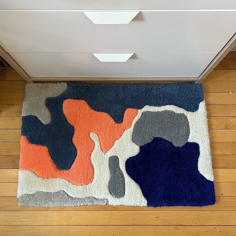 K&ouml;nnte beinhalten: Ein abstrakter Teppich in Wei&szlig;, Grau, Blau und Orange mit einem modernen, geometrischen Design. Der Teppich besteht aus flauschigem, strukturiertem Material.