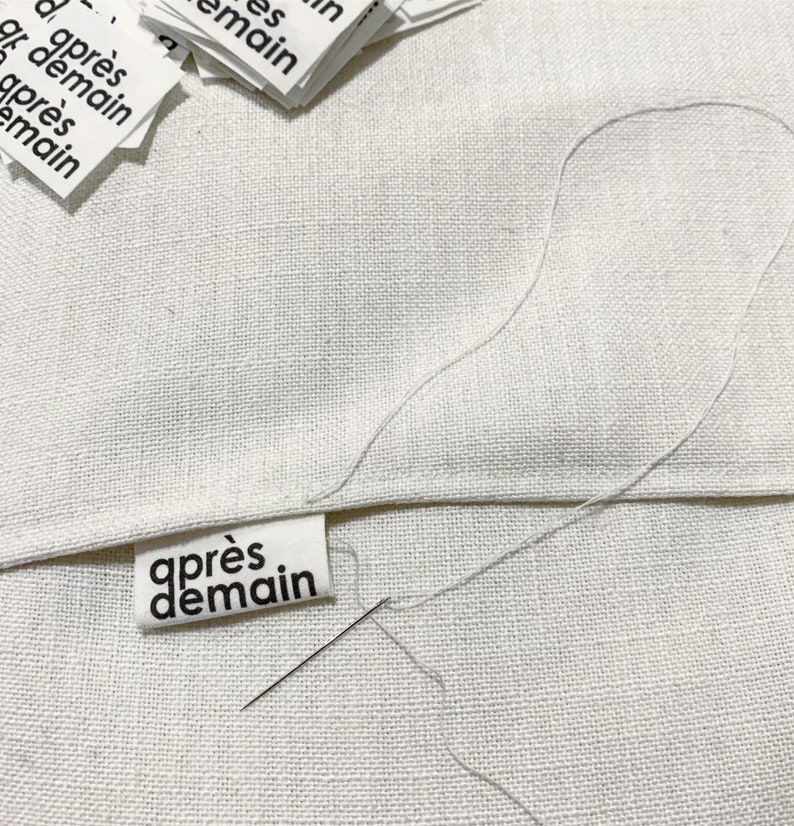 Op de afbeelding: Een wit stoffen label met zwarte tekst die "apr&egrave;s demain" luidt, is genaaid op een wit stoffen oppervlak. Een naald en witte draad zijn zichtbaar.