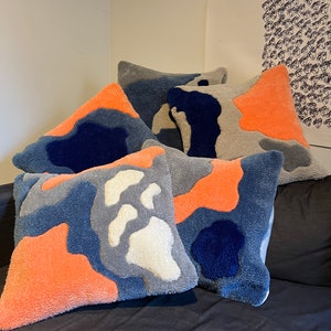 Op de afbeelding: Vijf decoratieve kussens met een kleurrijk abstract patroon in tinten blauw, grijs, wit en oranje. De kussens zijn gemaakt van een zachte, pluche stof en hebben een uniek, modern ontwerp.
