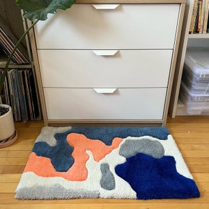 K&ouml;nnte beinhalten: Eine wei&szlig;e Kommode mit drei Schubladen und silbernen Griffen. Die Kommode steht vor einem Teppich mit einem abstrakten Design in Blau, Grau und Orange.